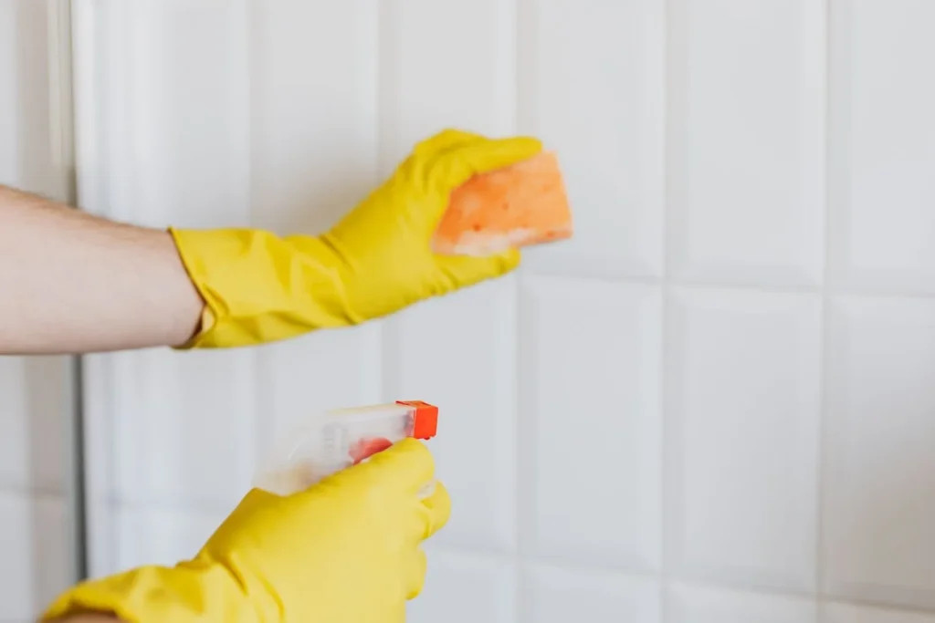 Gloved hands spraying and wiping bathroom tiles during mold and mildew removal